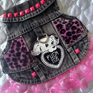May include: A dark gray acid-wash denim pet vest featuring a pink leopard print fabric on the chest panels, a row of neon pink square studs on the collar, and a central patch of a dog holding a heart with the text FUCK ICE and pink lace trim.