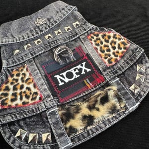 May include: A gray denim dog vest with leopard print patches and silver pyramid studs. The vest features a patch with the word "NOFX" and a skull emblem. A metal ring is attached for leash use.