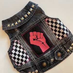 May include: A black denim vest with silver studs, a pink fist graphic, and black and white checkered fabric patches.