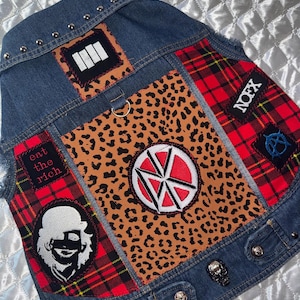 May include: A blue denim vest featuring a central leopard print panel, red and black plaid side panels, and various patches including a white and red circular logo, a black and white figure, and text reading eat the rich and NOFX.