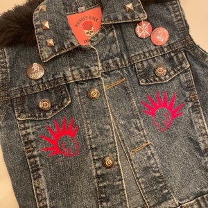 May include: A blue denim vest with a black faux fur collar, silver studs, and red punk rock patches. The vest has two pockets and a button closure. The patches include a skull with a rose, a punk rock symbol, and a logo that says "Whiskey Kale Crew".