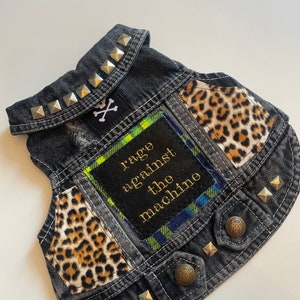 May include: A black denim vest with silver studs, a leopard print fabric patch, and a green and blue plaid patch with the text "rage against the machine" in gold lettering.