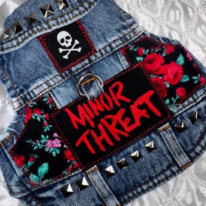 May include: A blue denim vest featuring silver pyramid studs along the collar and hem. It includes a black patch with a white skull and crossbones, a black patch with red roses, and a black patch with the red text MINOR THREAT and a metal D-ring.