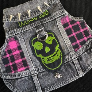 Puede incluir: Un chaleco vaquero gris con una estética punk rock. Cuenta con púas plateadas, un parche de calavera verde y detalles a cuadros rosas y negros. La palabra "Misfits" está bordada en verde.