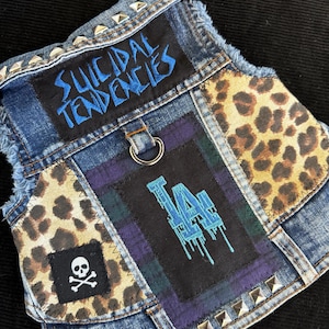 Könnte beinhalten: Eine Jeansweste mit Aufnähern und Metallnieten. Aufnäher mit dem Text "SUICIDAL TENDENCIES" in Blau, einem Totenkopf mit gekreuzten Knochen und "LA" in tropfendem Blau. Leopardenmuster und Schottenkaro-Akzente.