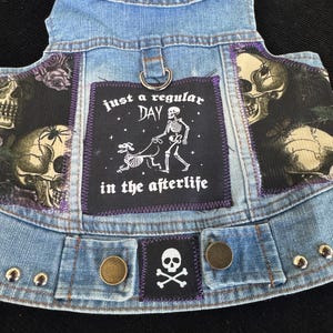 Puede incluir: Un chaleco vaquero azul claro para perros con parches. El chaleco presenta un parche con un esqueleto paseando a un perro y el texto "just a regular DAY in the afterlife". Parches adicionales incluyen calaveras y una calavera y huesos cruzados.