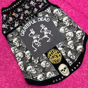 May include: Black sleeveless vest with a skull and rose pattern, featuring patches of the Grateful Dead, The Rolling Stones, and a skull with a top hat. The vest is decorated with silver studs.