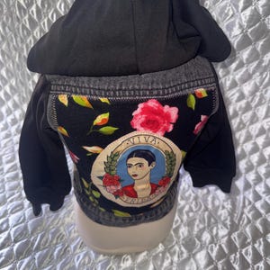 May include: A black hooded jacket featuring a dark denim back panel with a floral print and a circular patch in the centre. The patch displays an illustration of a person with dark hair and the text VIVA FRIDA in a decorative border.