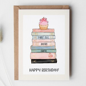 Pode incluir: Cartão de aniversário com uma pilha de livros em cores pastel e um cupcake rosa. As palavras "Here's to", "Another" e "Chapter" são visíveis. O texto "Happy Birthday!" está em baixo.
