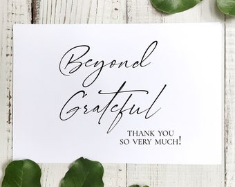 May include: White card with black calligraphy that reads "Beyond Grateful" and "Thank You So Very Much!" The card is on a white wood background with green leaves.