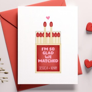 Puede incluir: Una tarjeta de San Valentín con un sobre rojo. La tarjeta presenta una ilustración de una caja de cerillas con dos cerillas sonrientes y el texto "I'M SO GLAD WE MATCHED" y los nombres "JESSICA + HENRY". También se ven dos decoraciones rojas en forma de corazón.