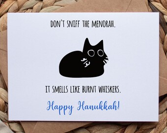 May include: A white Hanukkah greeting card with the text "DON'T SNIFF THE MENORAH. IT SMELLS LIKE BURNT WHISKERS. Happy Hanukkah!" and a black cat illustration. The card is on a brown envelope, set against a woven background.