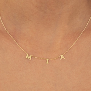 May include: A gold-colored necklace with the letters "MIA" displayed. The letters are individual and spaced apart. The necklace is worn against a red background.