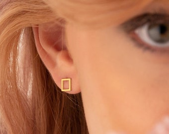 May include: A close-up of a woman's ear with a small gold square earring. The earring is simple and minimalist, with a thin, delicate frame.