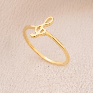May include: A delicate gold ring featuring a treble clef design. The ring is a thin band with the musical symbol as the focal point. The ring is set against a soft, neutral background, with a gold chain visible in the upper right corner.