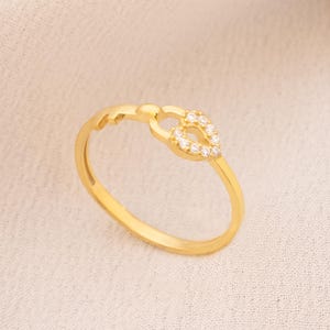 May include: A gold ring with a heart-shaped design. The heart is adorned with small, clear gemstones. The ring is set against a soft, off-white background, with a gold chain visible in the upper right corner.