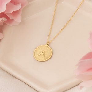 Op de afbeelding: Een gouden ketting met een ronde hanger met de letter 'A' in een geborstelde gouden afwerking.