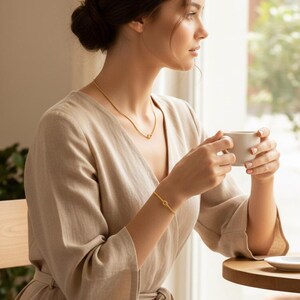 Op de afbeelding: Een gouden ketting- en armbandset. De ketting is een delicate ketting met een kleine cirkelvormige hanger. De armband heeft een vergelijkbaar ontwerp. De sieraden worden gedragen met een beige jurk. De persoon houdt een witte mok vast.