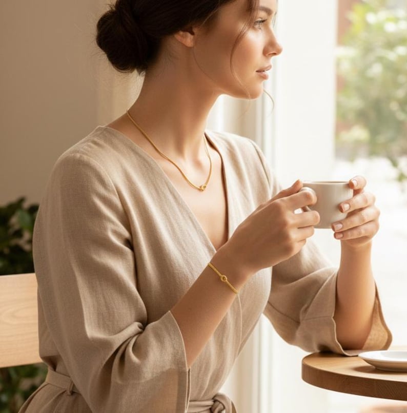 Op de afbeelding: Een gouden ketting- en armbandset. De ketting is een delicate ketting met een kleine cirkelvormige hanger. De armband heeft een vergelijkbaar ontwerp. De sieraden worden gedragen met een beige jurk. De persoon houdt een witte mok vast.