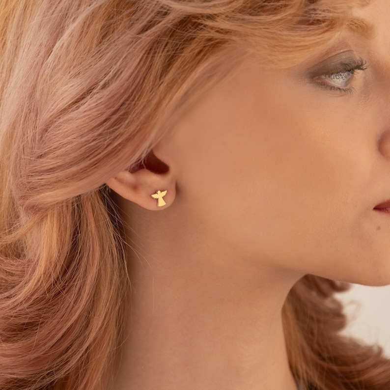 Puede incluir: Un par de pendientes de bot&oacute;n dorados con forma de &aacute;ngel. Los pendientes son peque&ntilde;os, con una silueta sencilla de un &aacute;ngel. Los pendientes se llevan en la oreja de una persona con piel clara y cabello rubio rojizo.