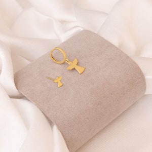 Puede incluir: Un conjunto de joyas de color dorado con un dise&ntilde;o de &aacute;ngel. El conjunto incluye un par de pendientes de aro con un colgante de &aacute;ngel y un solo pendiente de bot&oacute;n con un dise&ntilde;o de &aacute;ngel. Los pendientes se muestran sobre una superficie gris sobre un fondo blanco.
