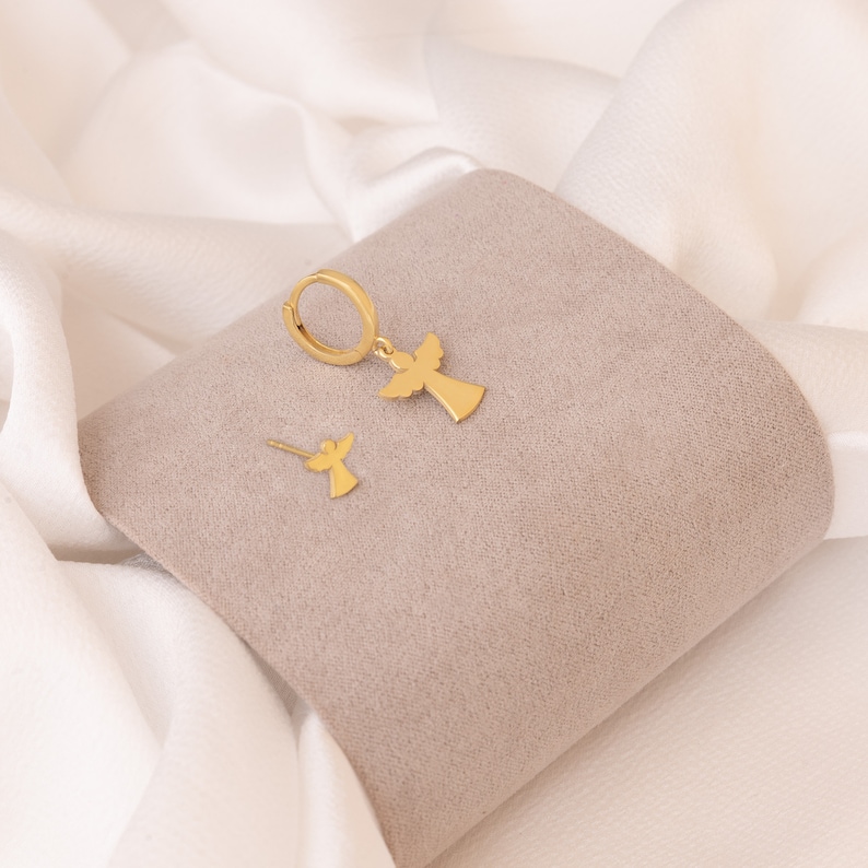 Puede incluir: Un conjunto de joyas de color dorado con un dise&ntilde;o de &aacute;ngel. El conjunto incluye un par de pendientes de aro con un colgante de &aacute;ngel y un solo pendiente de bot&oacute;n con un dise&ntilde;o de &aacute;ngel. Los pendientes se muestran sobre una superficie gris sobre un fondo blanco.
