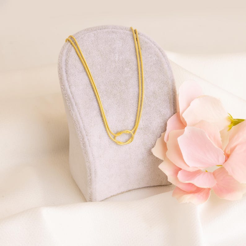Op de afbeelding: Een goudkleurige ketting met een dubbelstrengs ontwerp, gepresenteerd op een grijze sieradenstandaard. De ketting heeft een knoopvormige hanger. Een roze bloem staat op de achtergrond, tegen een witte stof.