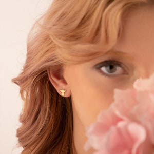 Puede incluir: Primer plano de una oreja con un pendiente de &aacute;ngel dorado. El pendiente tiene forma de un peque&ntilde;o &aacute;ngel estilizado. La persona tiene la piel clara, el pelo rojo claro y una flor rosa que le cubre parcialmente la cara.