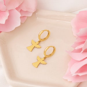Puede incluir: Un par de pendientes de aro dorados con colgantes en forma de &aacute;ngel. Los pendientes se muestran sobre una superficie blanca, con p&eacute;talos de flores rosas en el fondo. Los colgantes de &aacute;ngel tienen alas extendidas y una silueta sencilla.