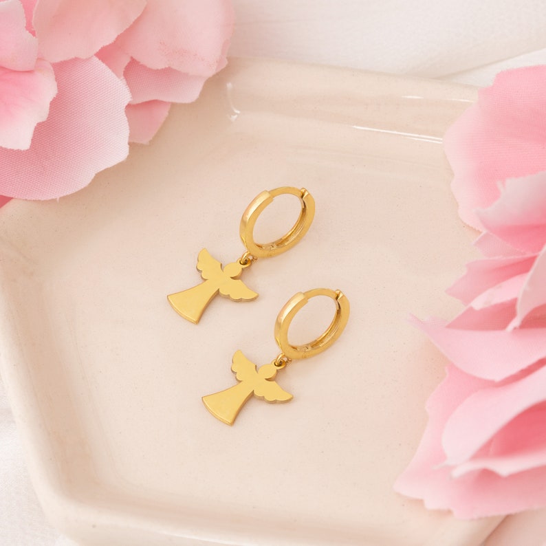 Puede incluir: Un par de pendientes de aro dorados con colgantes en forma de &aacute;ngel. Los pendientes se muestran sobre una superficie blanca, con p&eacute;talos de flores rosas en el fondo. Los colgantes de &aacute;ngel tienen alas extendidas y una silueta sencilla.