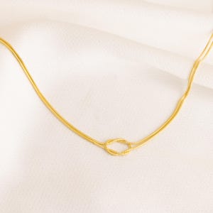 Op de afbeelding: Een goudkleurige ketting met een knoopontwerp. De ketting heeft een delicate slangenkettingstijl en wordt gepresenteerd op een witte, getextureerde ondergrond. De knoop is het middelpunt van de ketting.