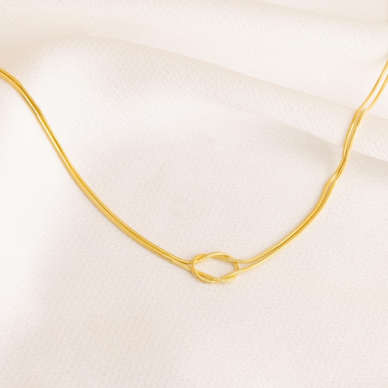 Op de afbeelding: Een goudkleurige ketting met een knoopontwerp. De ketting heeft een delicate slangenkettingstijl en wordt gepresenteerd op een witte, getextureerde ondergrond. De knoop is het middelpunt van de ketting.