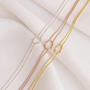 Op de afbeelding: Drie delicate kettingen in zilver, ros&eacute;goud en goud, elk met een knoopontwerp. De kettingen worden gepresenteerd op een witte, getextureerde ondergrond, die hun strakke en minimalistische stijl benadrukt. De kettingen zijn dun en elegant.