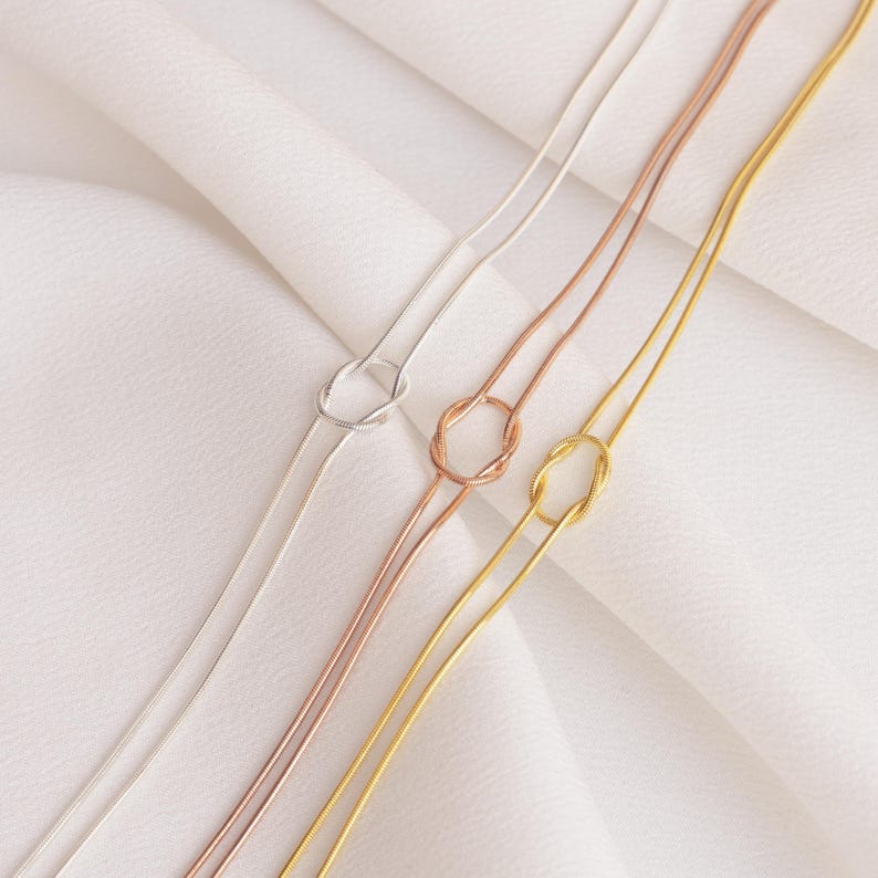 Op de afbeelding: Drie delicate kettingen in zilver, ros&eacute;goud en goud, elk met een knoopontwerp. De kettingen worden gepresenteerd op een witte, getextureerde ondergrond, die hun strakke en minimalistische stijl benadrukt. De kettingen zijn dun en elegant.