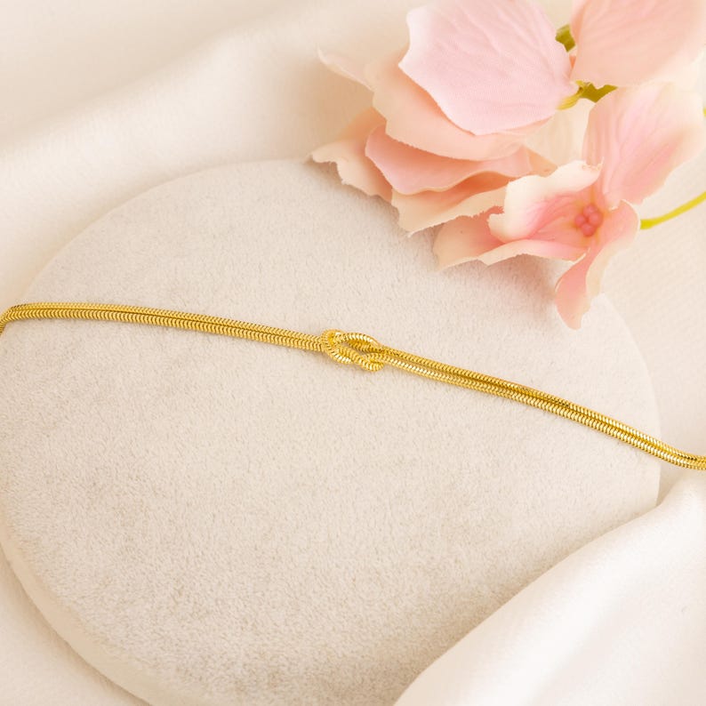 Op de afbeelding: Een goudkleurige armband met een knoopontwerp. De armband is gemaakt van een dunne, slangachtige ketting. De achtergrond omvat een wit oppervlak en een roze bloem.