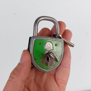 Puede incluir: Un candado vintage verde y plateado con un ojo de cerradura y una llave adjunta. El candado tiene un diseño en forma de escudo con el texto "H.M. EURO" en la parte delantera.