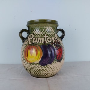 May include: A ceramic vase with a green rim and handles, featuring a textured beige body. The vase is decorated with a painted design of a pumpkin, eggplant, and strawberry, with the word "Rumtons" above. The vase is a decorative kitchen item.
