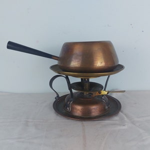 May include: A copper fondue pot with a black handle sits on a copper stand with a black metal base. The stand has a small burner for heating the pot.