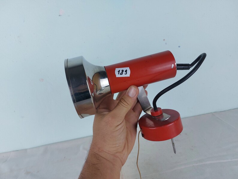 May include: A red and chrome wall-mounted lamp with a flexible arm and a round base. The lamp has a chrome reflector and a red cord. The lamp is labeled "189".