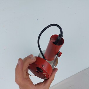 May include: A red and silver ceiling light fixture with a black wire. The light fixture is being held by a hand.