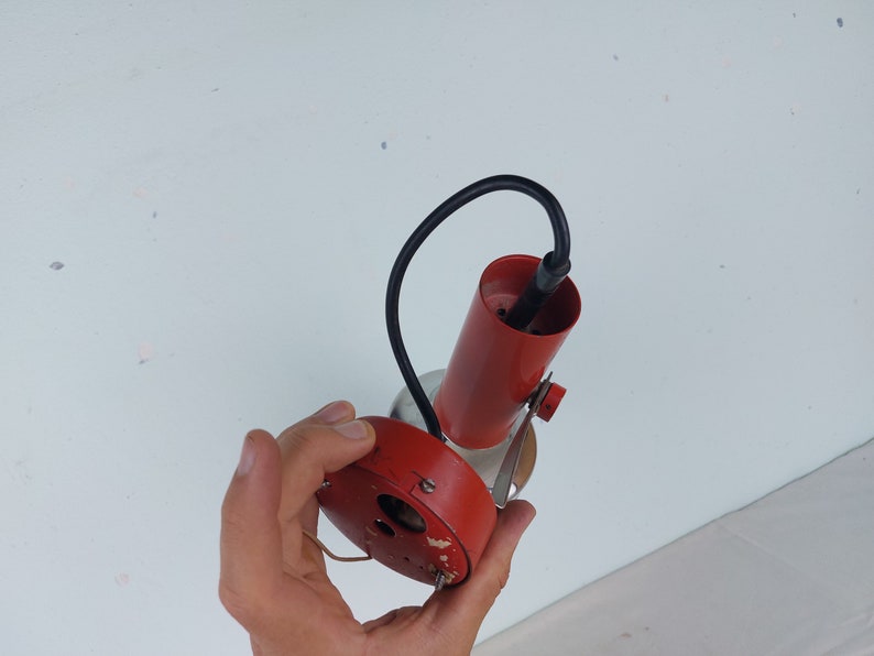 May include: A red and silver ceiling light fixture with a black wire. The light fixture is being held by a hand.