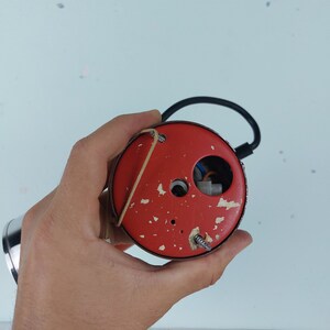 May include: Close-up of the back of a red round electrical outlet cover with two screws and a small hole in the center. The cover is held in a hand.