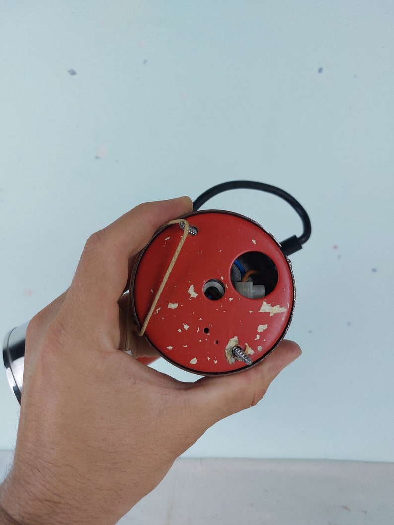 May include: Close-up of the back of a red round electrical outlet cover with two screws and a small hole in the center. The cover is held in a hand.