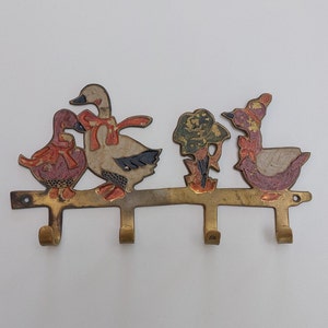 Puede incluir: Porta llaves de latón con cuatro ganchos con forma de patos. Los patos están pintados en tonos de rojo, naranja, blanco y negro. Los patos llevan cintas rojas y blancas. Un pequeño árbol verde y dorado se encuentra entre dos de los patos.