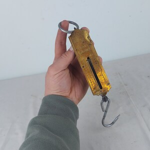 Könnte beinhalten: Eine antike Federwaage aus Messing mit Metallhaken und Ring. Die Waage hat ein vergilbtes, gealtertes Aussehen mit sichtbaren Messmarkierungen. Die Waage wird in einer Hand vor weißem Hintergrund gehalten.
