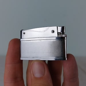 Puede incluir: Un encendedor plateado con una superficie texturizada y una pequeña perilla en el lateral.