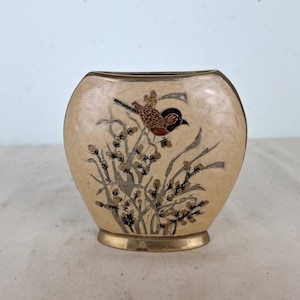 Puede incluir: Un jarrón pequeño de cerámica de forma ovalada, de color crema, con un borde dorado y un diseño pintado de un pájaro y flores.