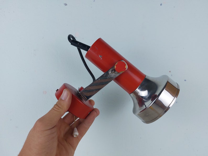 May include: A red and chrome adjustable wall lamp with a black cord. The lamp has a round base and a long, thin arm that can be moved up and down.