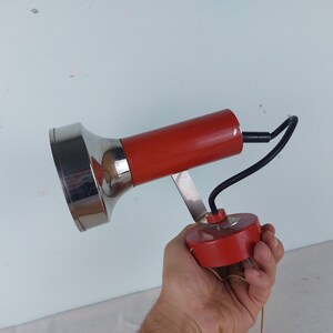 May include: A red and silver wall mounted lamp with a flexible arm. The lamp has a round base and a cylindrical shade.