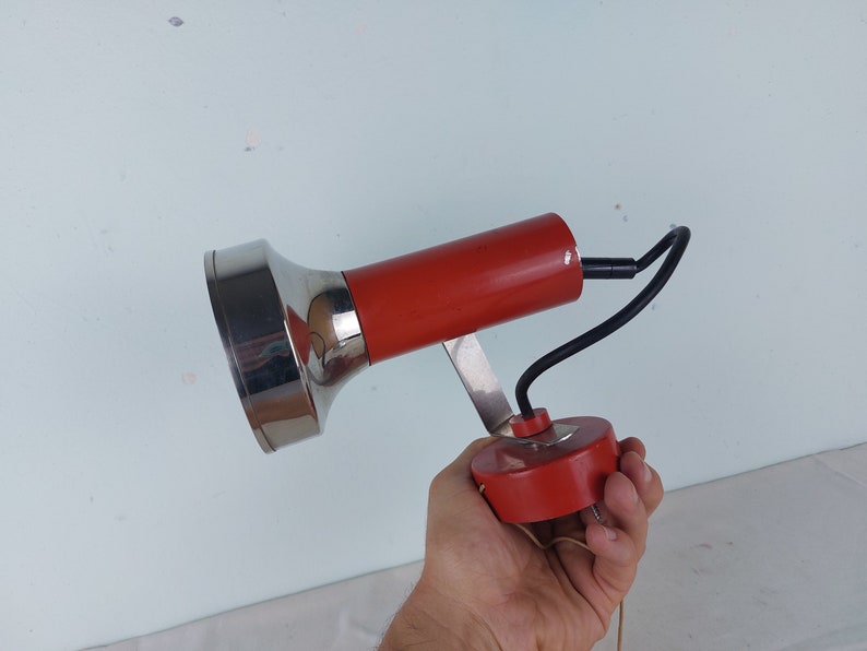 May include: A red and silver wall mounted lamp with a flexible arm. The lamp has a round base and a cylindrical shade.